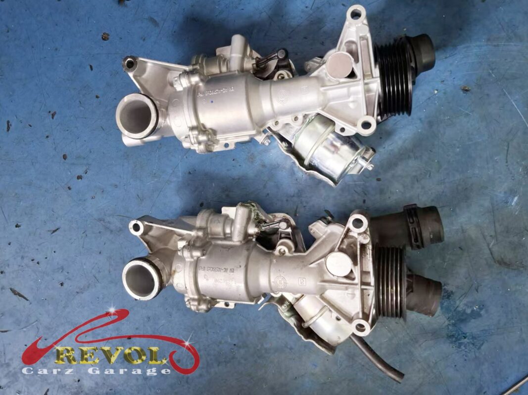 The old water pump assembly (bottom) beside the new Mercedes-spec unit (top), with the thermostat housing and pulley built in.
