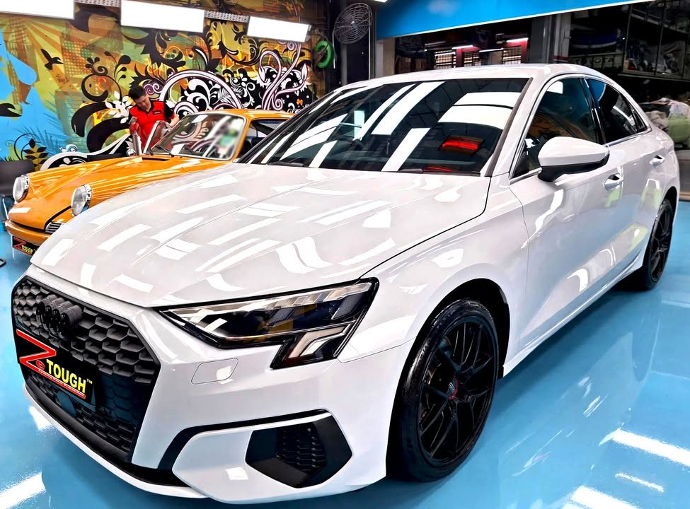 White Audi A3 sedan from the front three-quarter in the Revol workshop, with black wheels and a ZeTough plate on the grille.