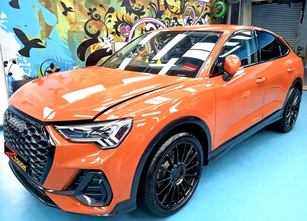 Orange Audi Q3, front three-quarter view, with black wheels and workshop lights reflected on the bonnet.