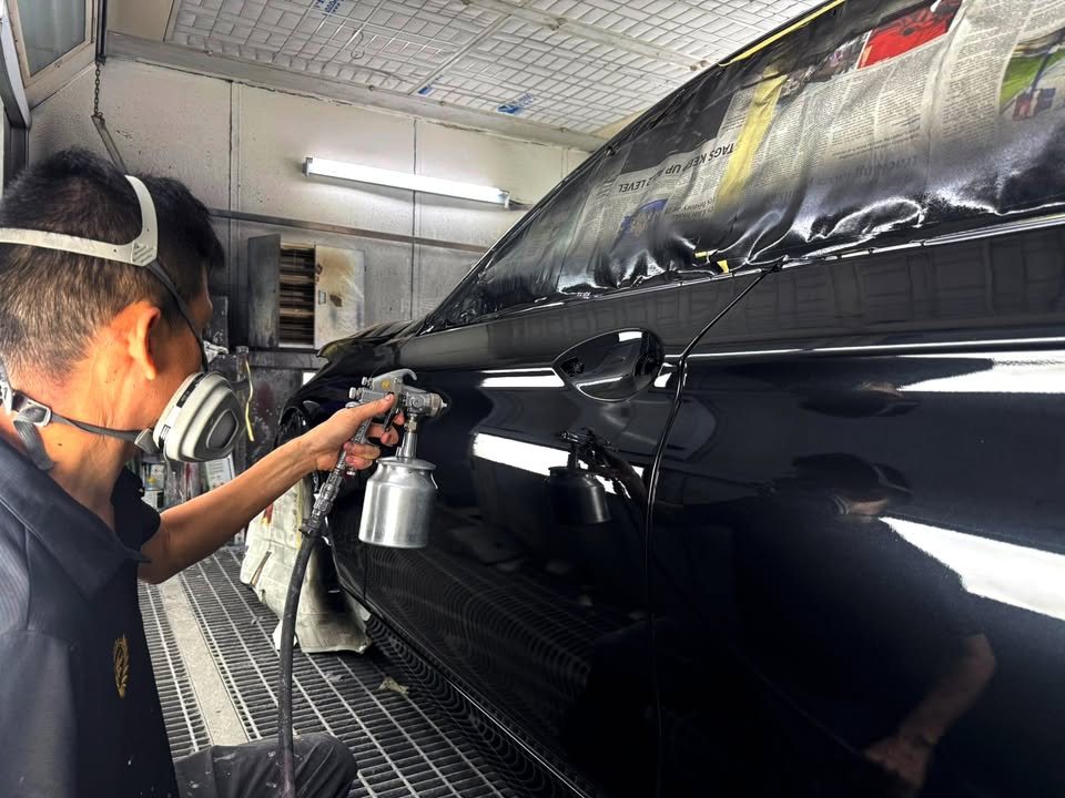 A painter in a respirator spraying the side of the black BMW 520i inside the spray booth, windows masked with paper.