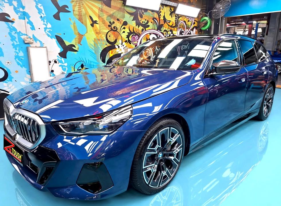 Front three-quarter view of a blue BMW i5 in the Revol Carz Makeover studio, with a ZeTough plate on the front bumper.