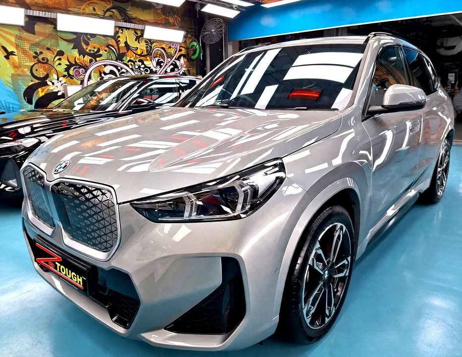 Front three-quarter view of a silver BMW iX1 in the Revol Carz Makeover studio, with a ZeTough plate on the front bumper.