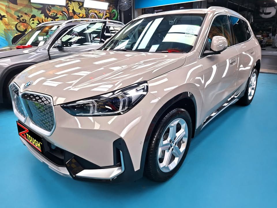 Beige BMW iX1, front three-quarter view in the workshop with a ZeTough plate on the bumper.