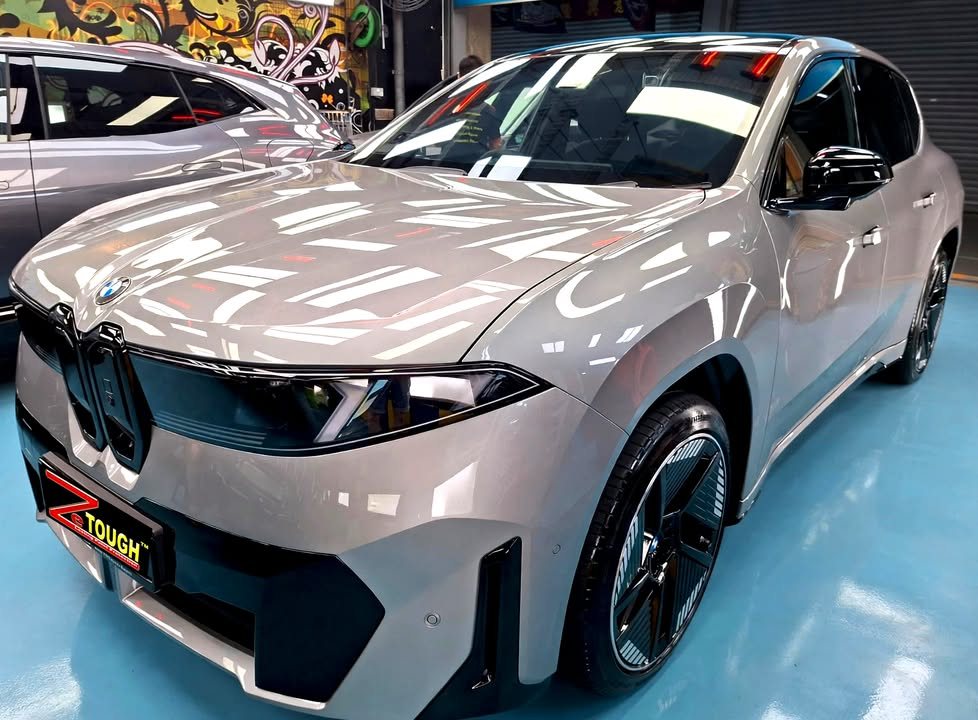 Grey BMW iX3, front three-quarter view in the workshop, with ceiling lights reflected in the bonnet and a ZeTough plate on the front.