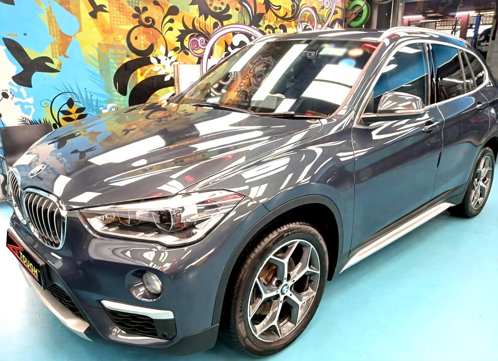 Front three-quarter view of a blue BMW X1 in the Revol Carz hall, with lights reflected across the bonnet.