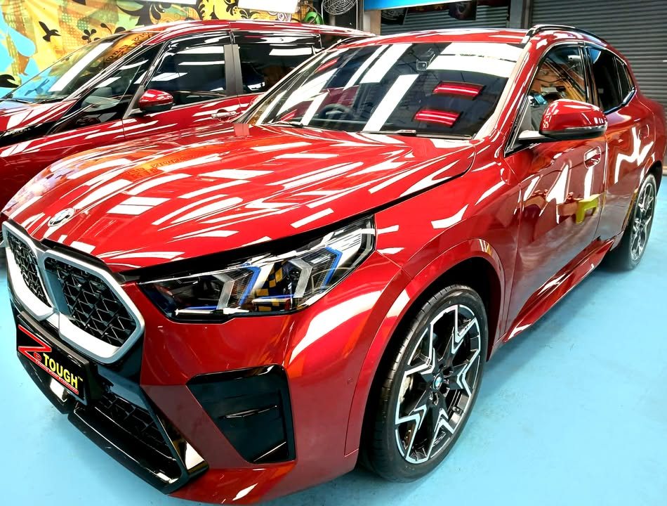 Front three-quarter view of a red BMW X2 in the Revol Carz hall, ceiling lights reflected across the bonnet.