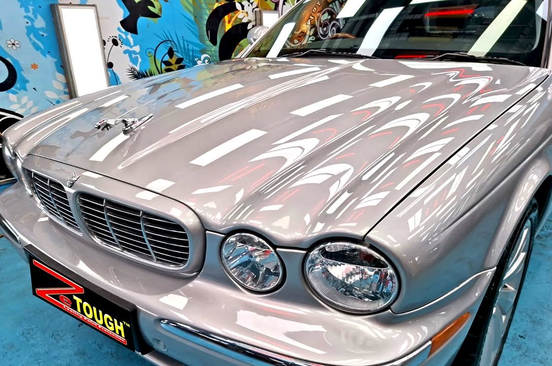 Front grille, twin round headlights and bonnet of the silver Jaguar XJ8 reflecting the workshop lights.