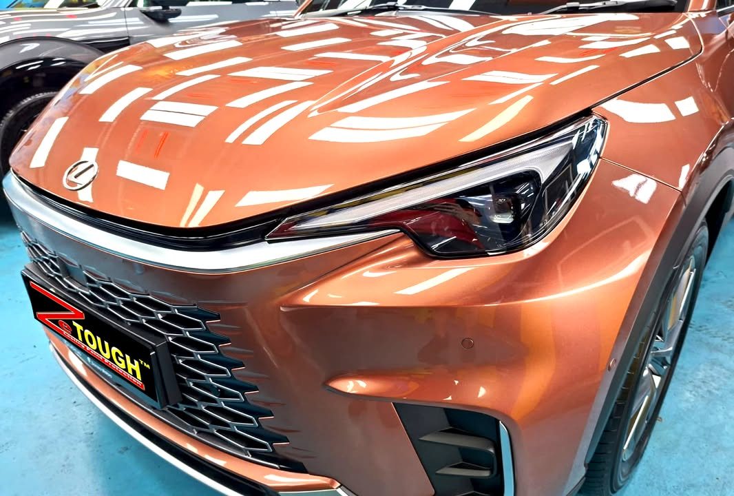 Front of the copper Lexus LBX showing the headlamp, grille and bonnet reflections.