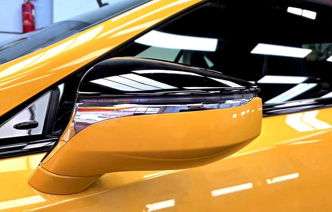Yellow side mirror with a black cap, ceiling lights reflected in the paint.