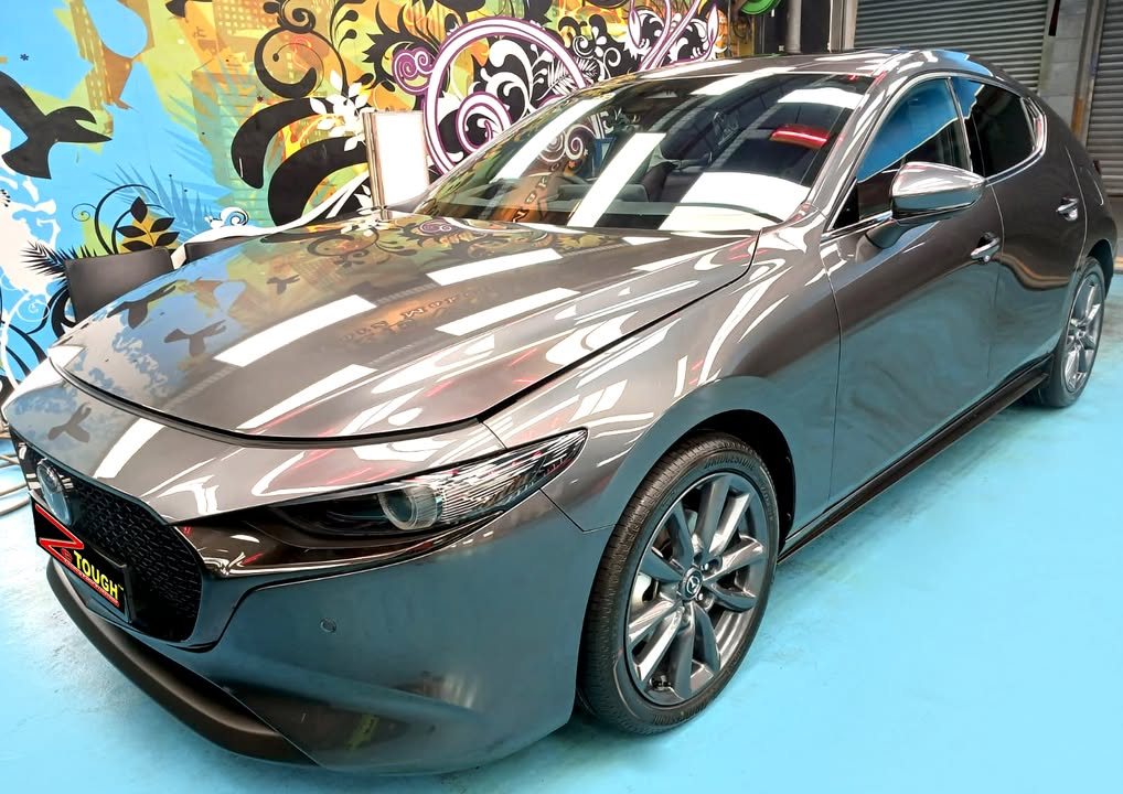 Grey Mazda 3 hatchback, front three-quarter view in the workshop, with ceiling lights reflected across the bonnet.