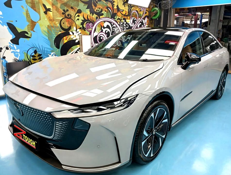 Front three-quarter view of a light grey Mazda 6e with a black roof in the Revol Carz Makeover studio, ZeTough plate on the bumper.