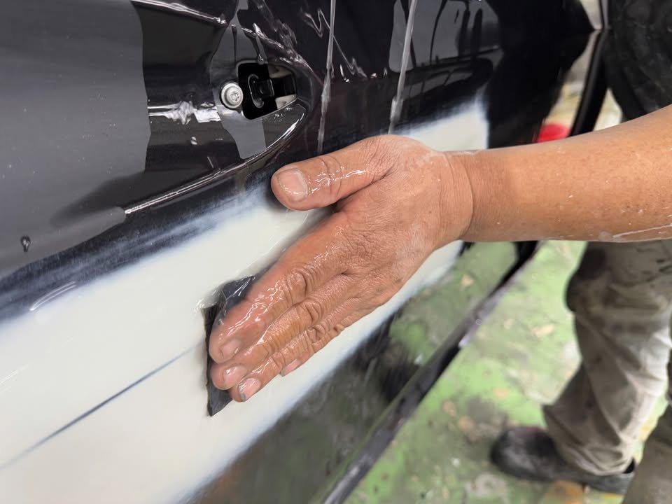 A hand wet-sanding the lower edge of a black door panel.