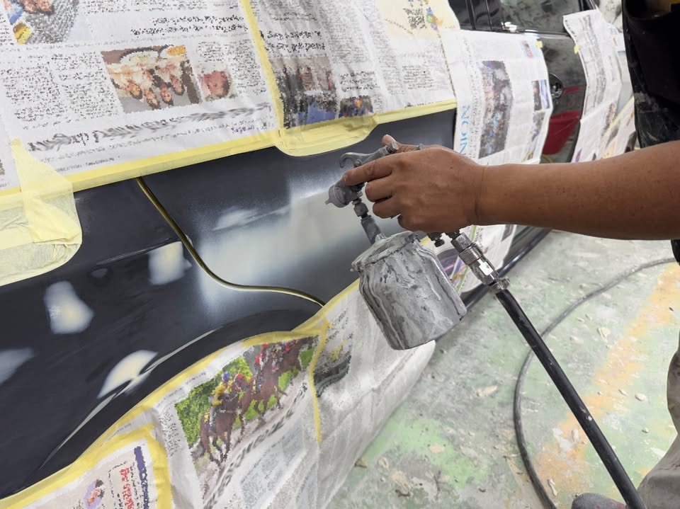 A spray gun applying black paint to a panel surrounded by newspaper masking.