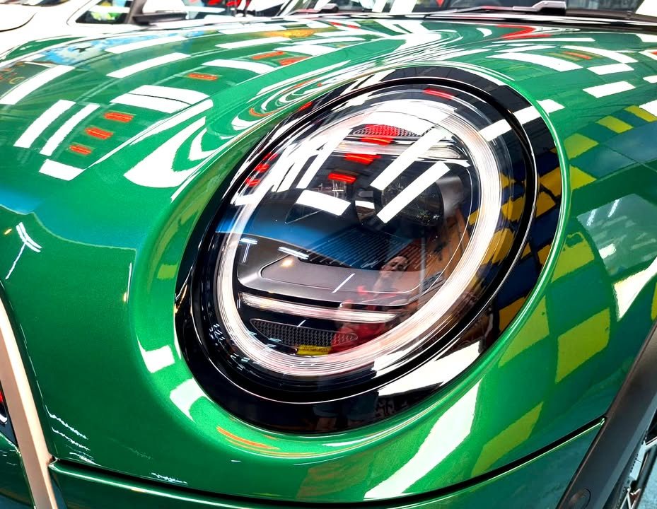 Close-up of the MINI Cooper's round headlamp with workshop lights reflected in the green paint.