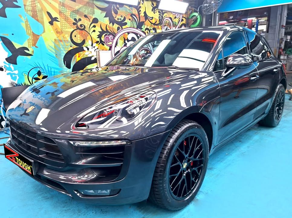 Dark grey Porsche Macan from the front three-quarter in the Revol workshop, with black wheels and a ZeTough plate.
