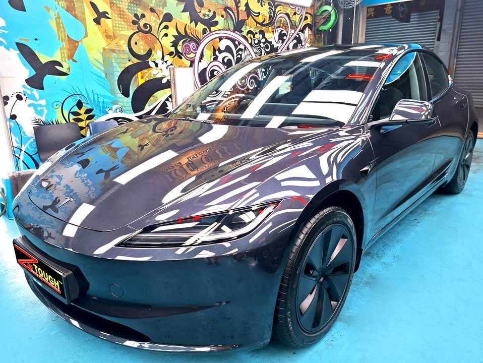 Dark grey Tesla Model 3 from the front three-quarter, workshop lights reflected across the coated bonnet.
