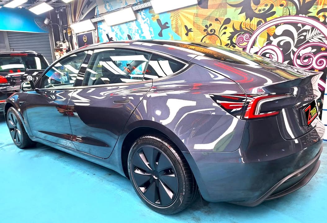 Rear three-quarter view of the grey Model 3 on the blue workshop floor.
