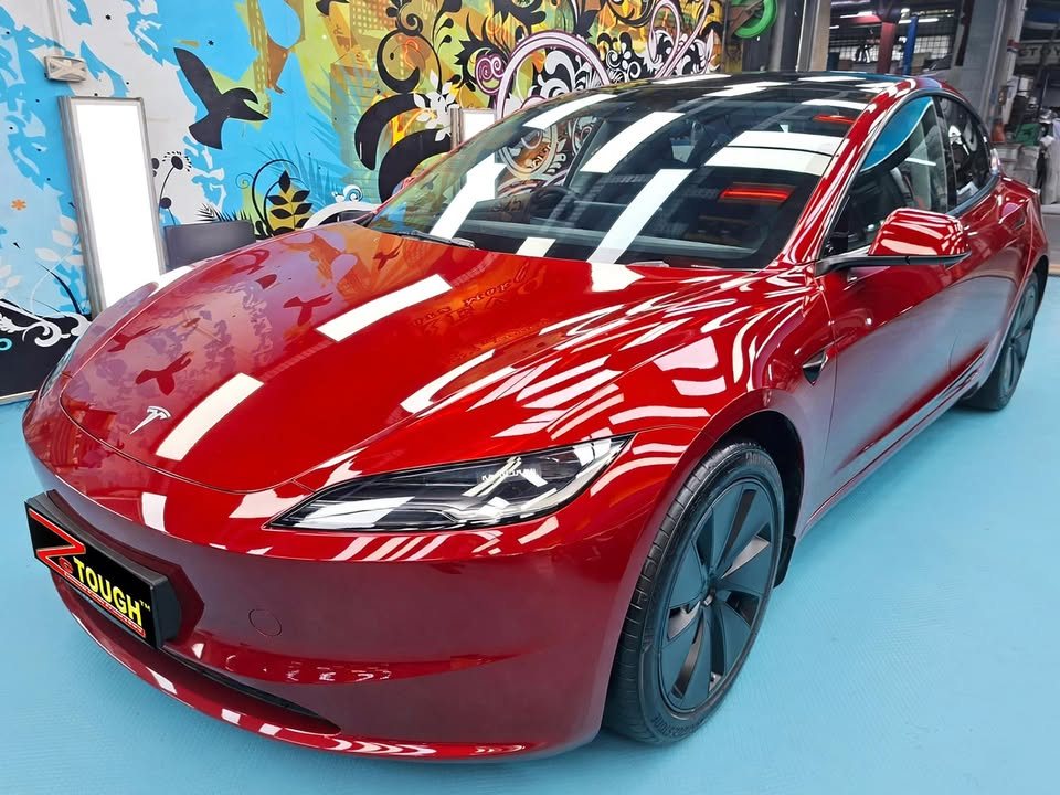 Front three-quarter view of the red Tesla Model 3 in the workshop, glossy paint reflecting the ceiling lights.