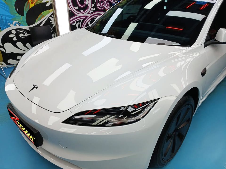 Front of the white Tesla Model 3 with the bonnet and slim headlamp reflecting the workshop lights.
