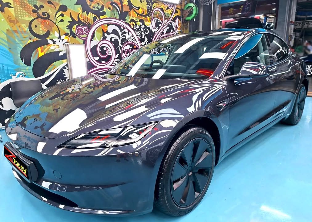 The dark grey Tesla Model 3 from the front three-quarter, the coated paint reflecting the workshop lights.