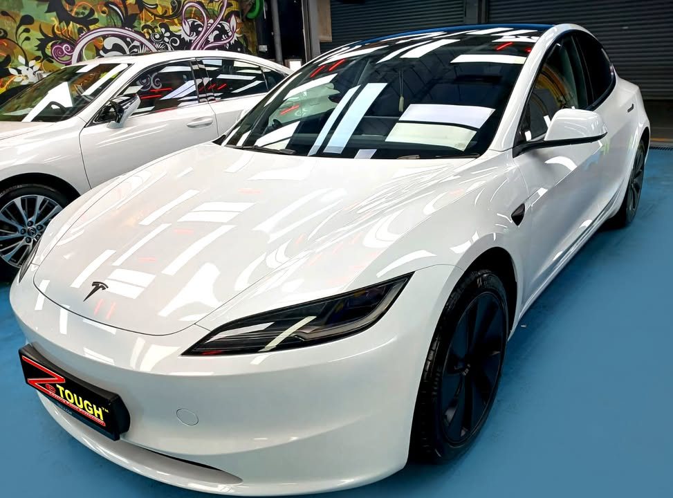 Front three-quarter view of the white Tesla Model 3 in the workshop, lights reflected on the bonnet.