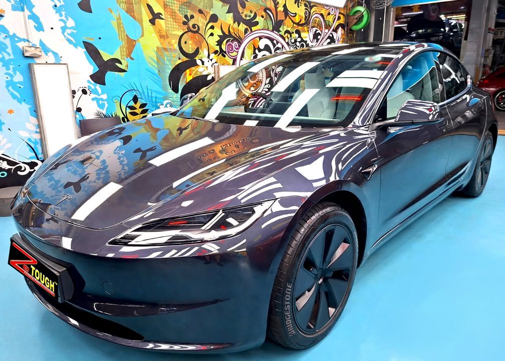 The dark grey Tesla Model 3 from the front three-quarter, its bonnet reflecting the workshop lights.