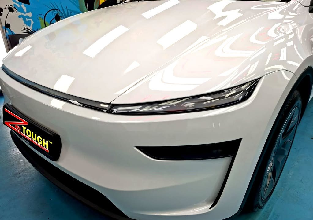 Front of the white Tesla Model Y L with its full-width light bar and bonnet reflecting the workshop lights.