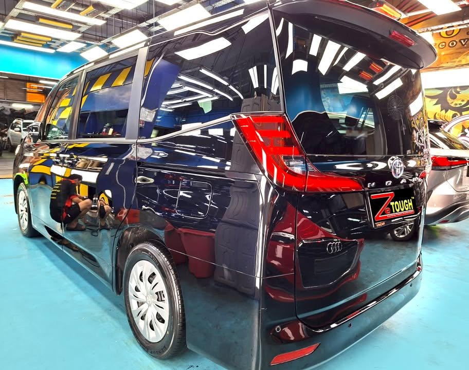 Rear three-quarter view of the black Toyota Noah with workshop lights reflected along its side.