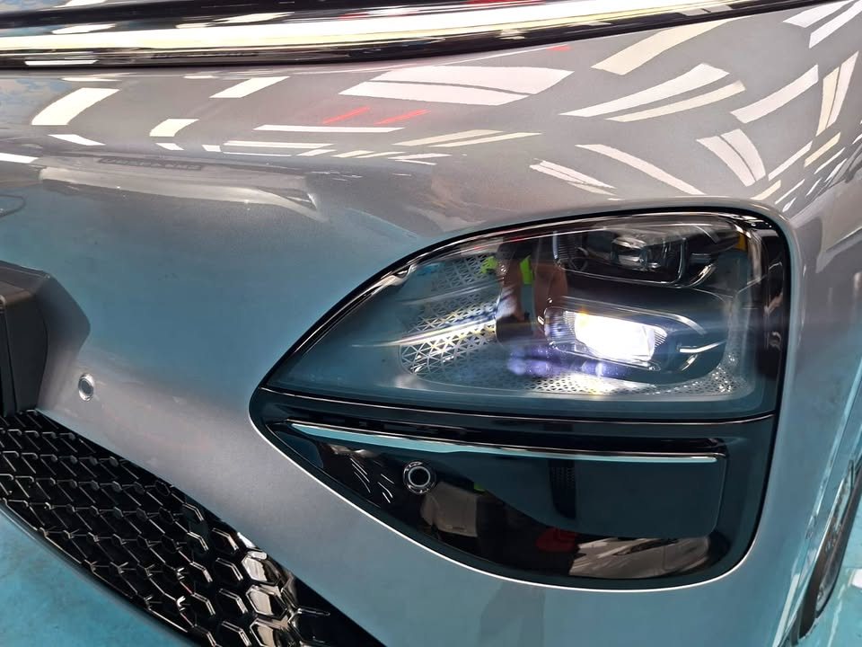 Close-up of the Xpeng G6 headlight and grey front bumper reflecting the workshop lights.