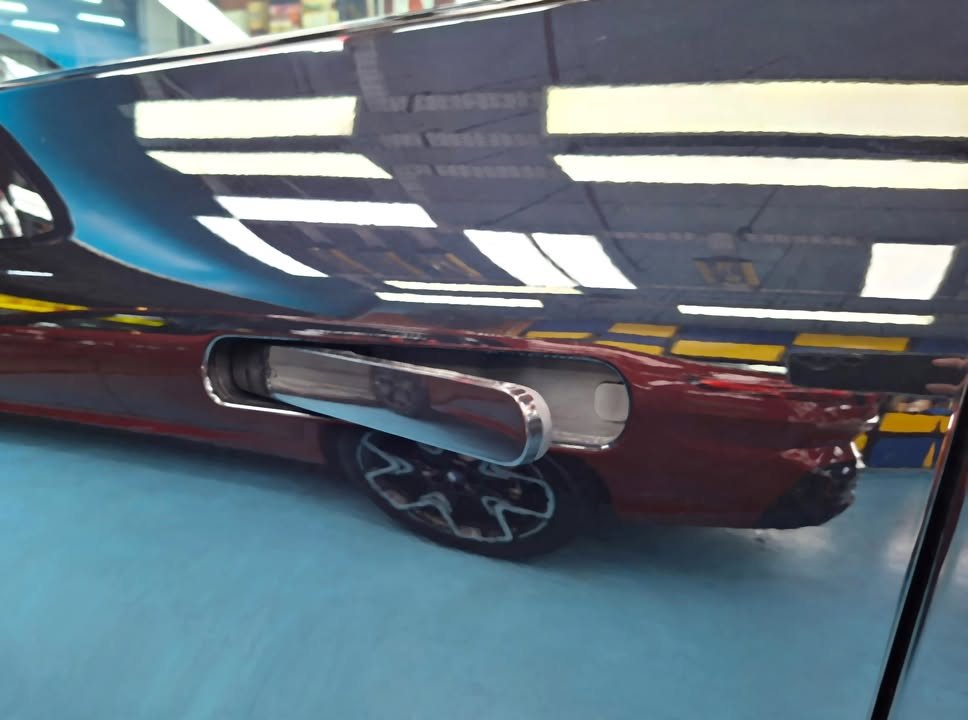 Flush door handle on a black door panel reflecting a red car parked alongside.