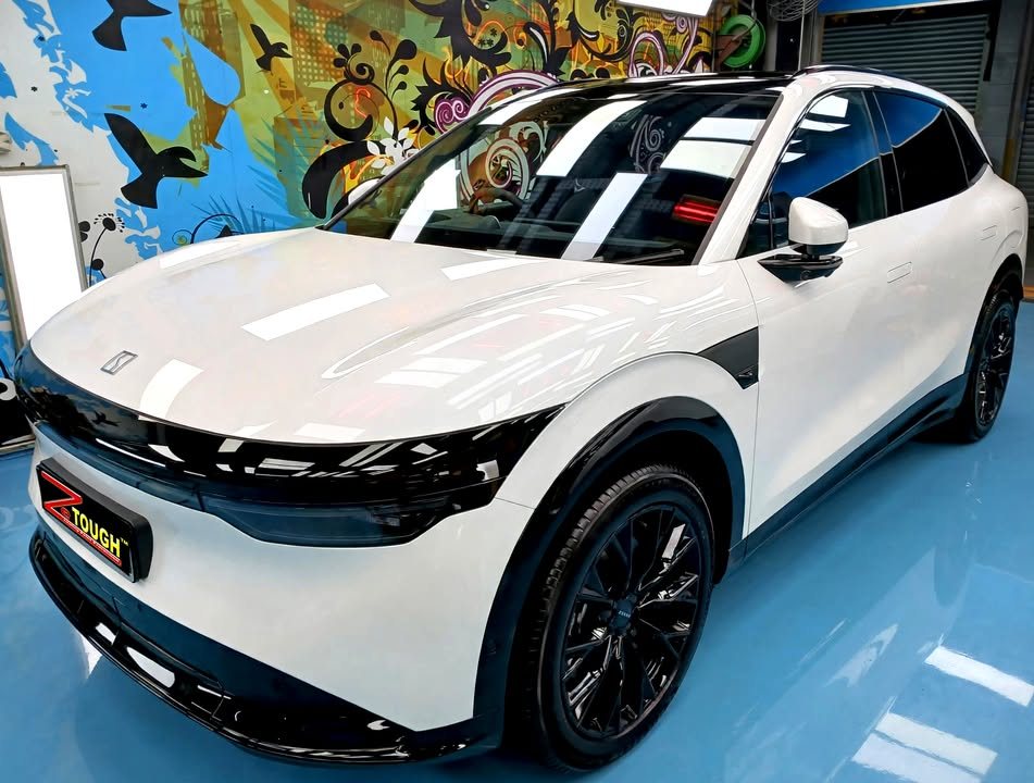 The white Zeekr 7X from the front three-quarter in the workshop, bonnet reflecting the ceiling lights.
